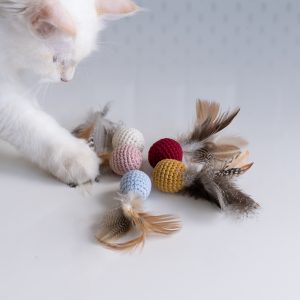 Kulka Pierzulka dla kota z kocmiętką i walerianą, kilka kolorów na białym stole, po lewo biały kot dotyka łapką zabawki