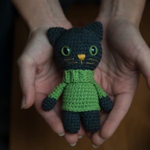 Szydełkowa maskotka czarny kot z zielonymi oczami w zielonym swetrze trzymana na dłoniach